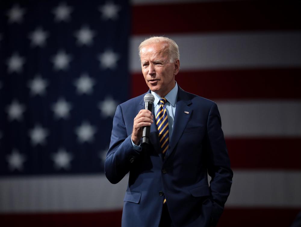 Joe Biden standing in front of an American flag, holding a microphone